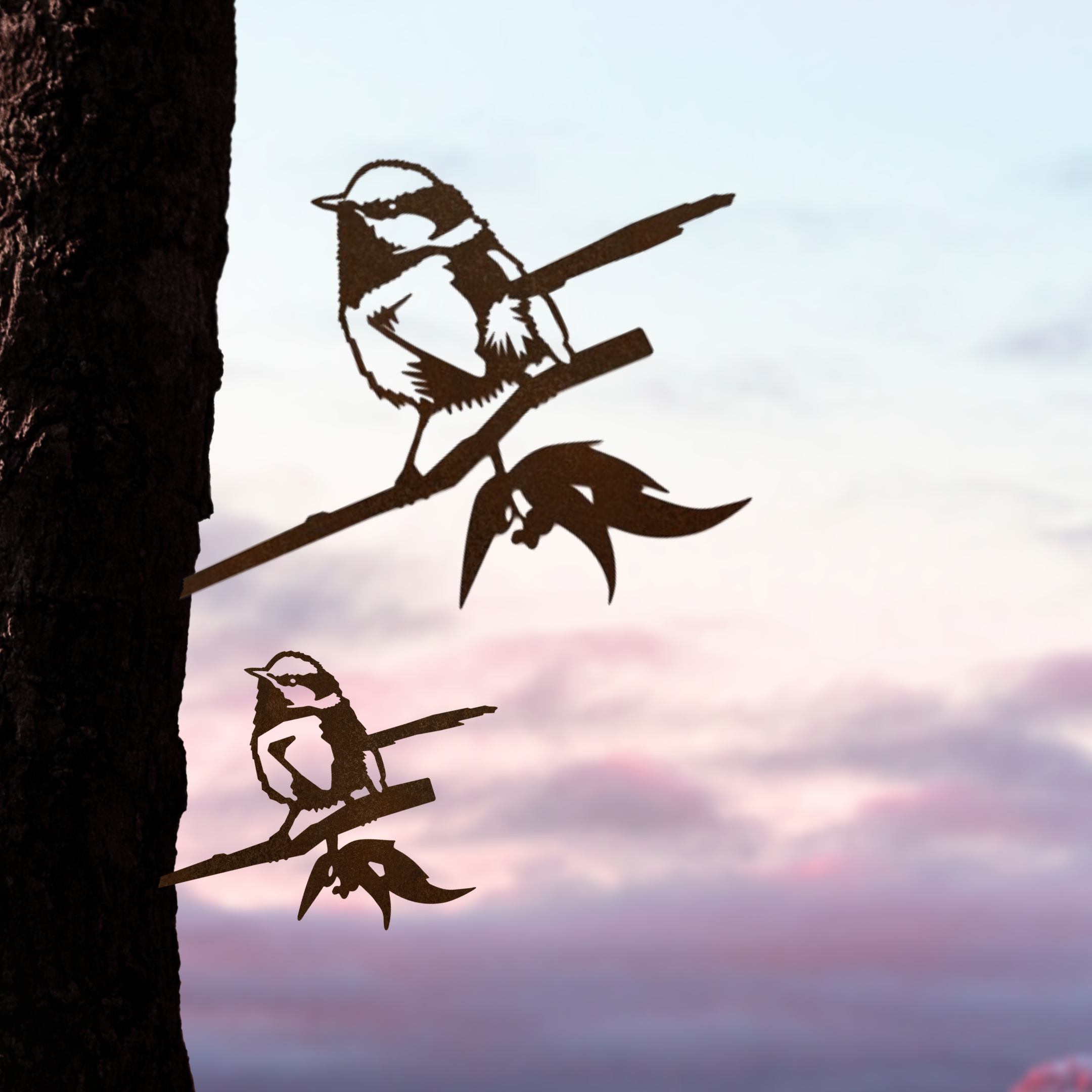 The Metalbird Australia Blue Wren Mum & Mini Set features two premium metal bird silhouettes on branches, attached to a tree trunk—an elegant yard art addition perfect against a pastel sunrise or sunset sky.