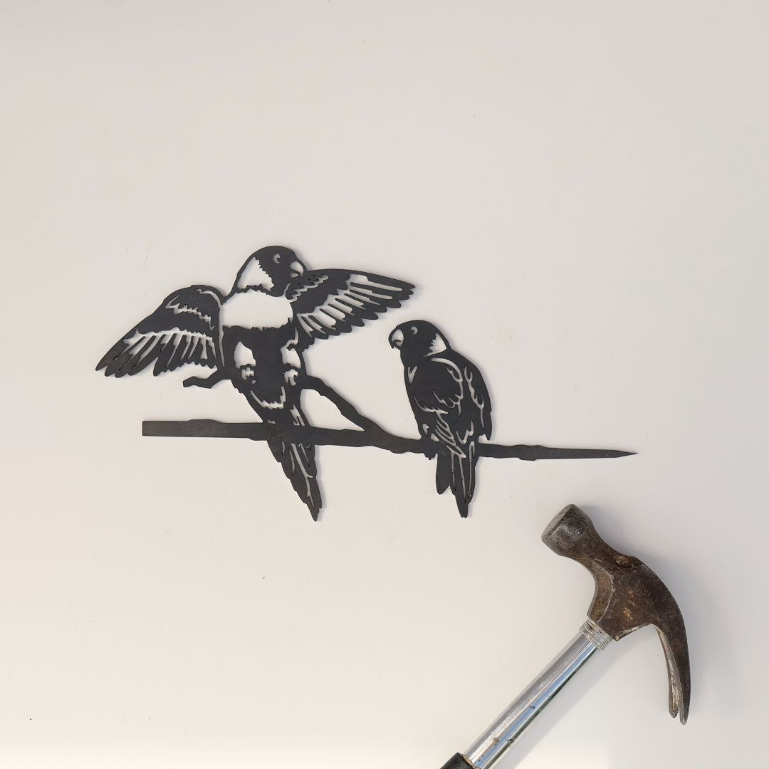The Pair of Lorikeets by Metalbird Australia features a corten steel silhouette of two birds on a branch, mounted on a white wall with a hammer below—suggesting recent installation. A charming and thoughtful gift idea.