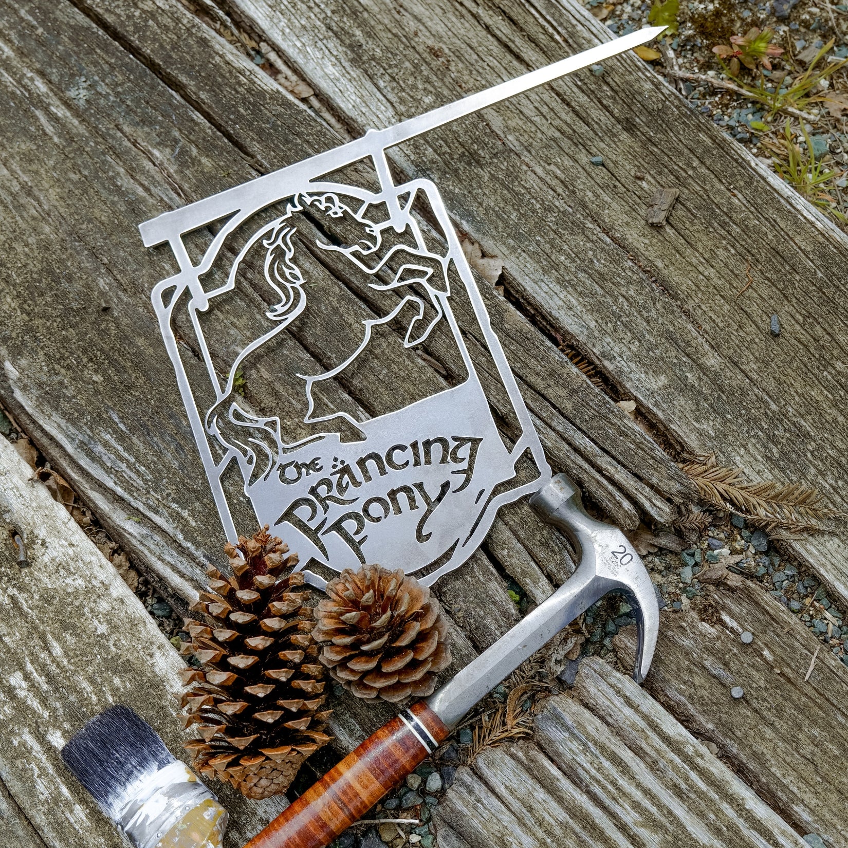 A premium THE PRANCING PONY™ metal sign by Metalbird Australia, featuring a prancing horse on weathered wood beside tools and pinecones—a perfect gift for fans.
