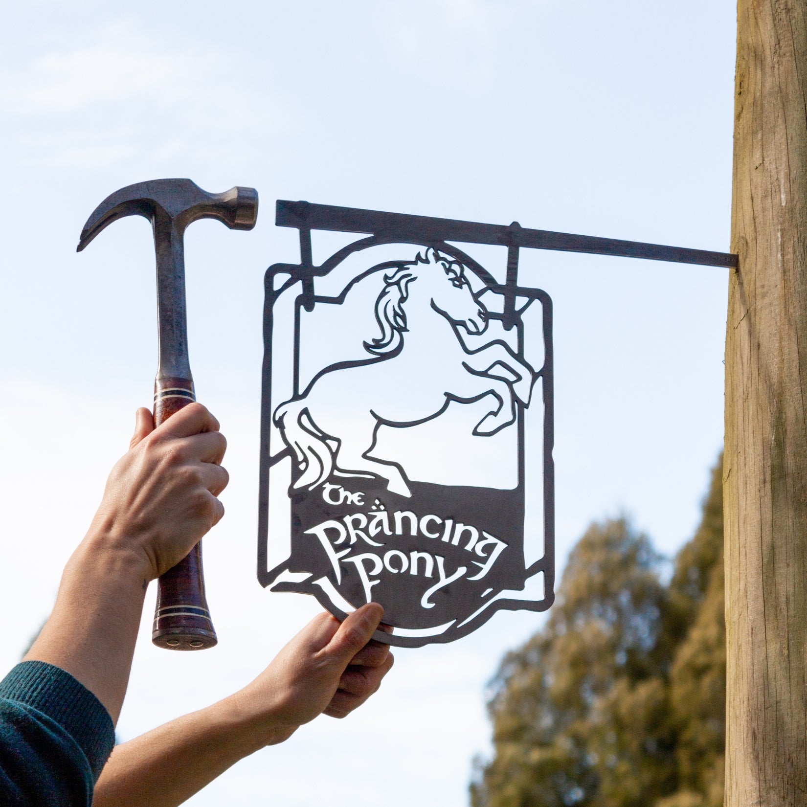 A person holds a Metalbird Australia metal sign featuring THE PRANCING PONY™ rearing horse silhouette, while another hand with a hammer is near a wooden post outdoors.