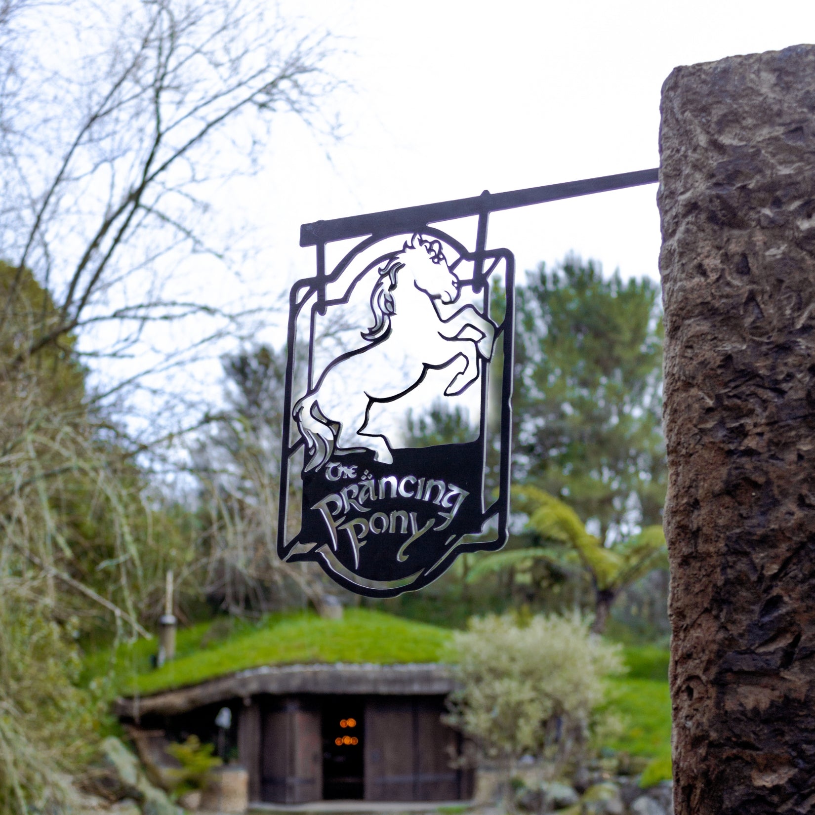 A premium metal sign by Metalbird Australia features THE PRANCING PONY™ silhouette and text, hanging from a stone post with a green hillside and rustic wooden building behind—an ideal gift for fans.