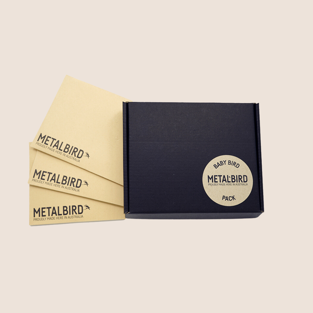 A premium black box with a round Baby Bird Bundle label sits beside three light brown envelopes marked Metalbird Australia—an ideal gift—on a plain beige background.