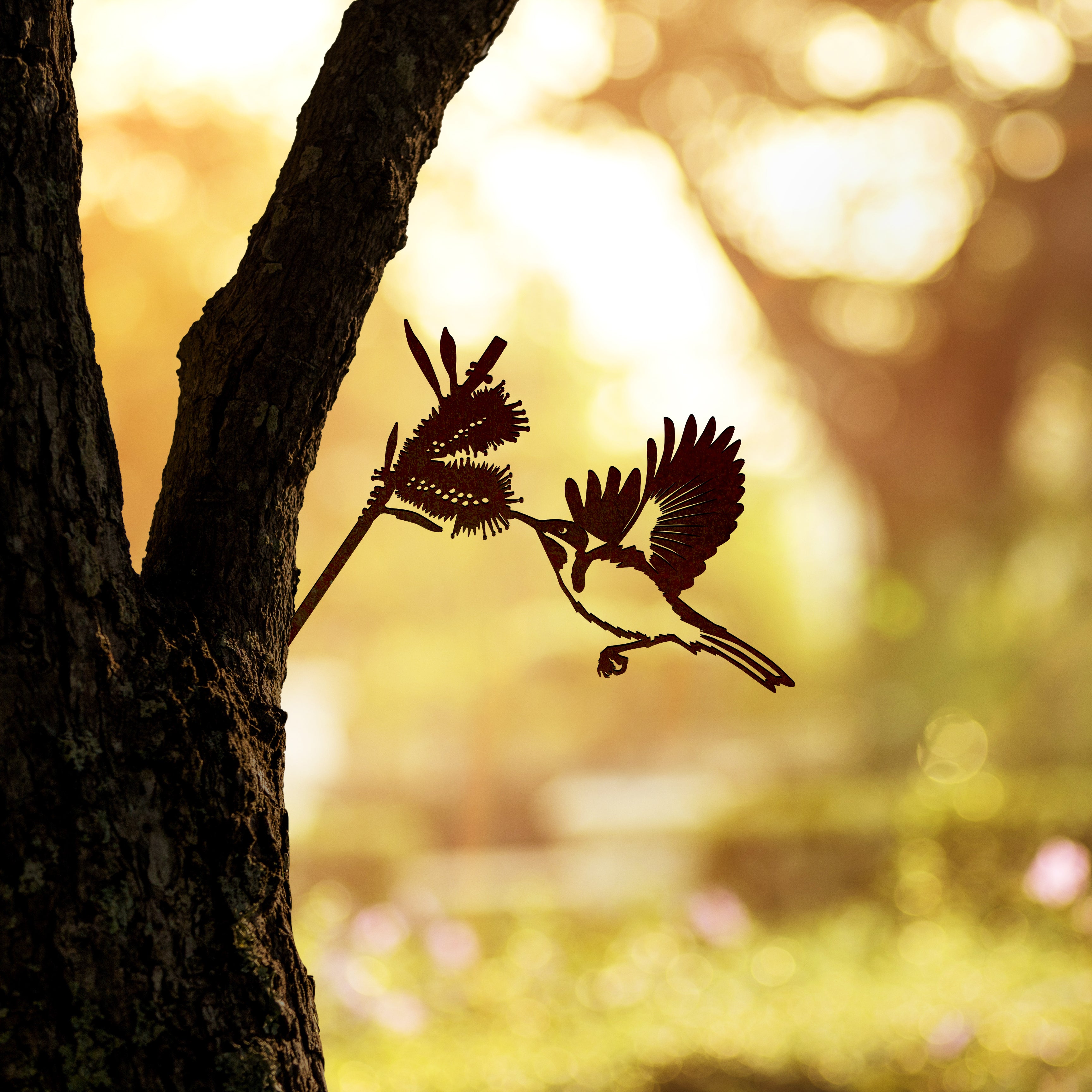 The Metalbird Australia Eastern Spinebill is a premium corten steel bird silhouette mounted on a tree, designed to look like it's perched among leaves, set against a sunlit green background.