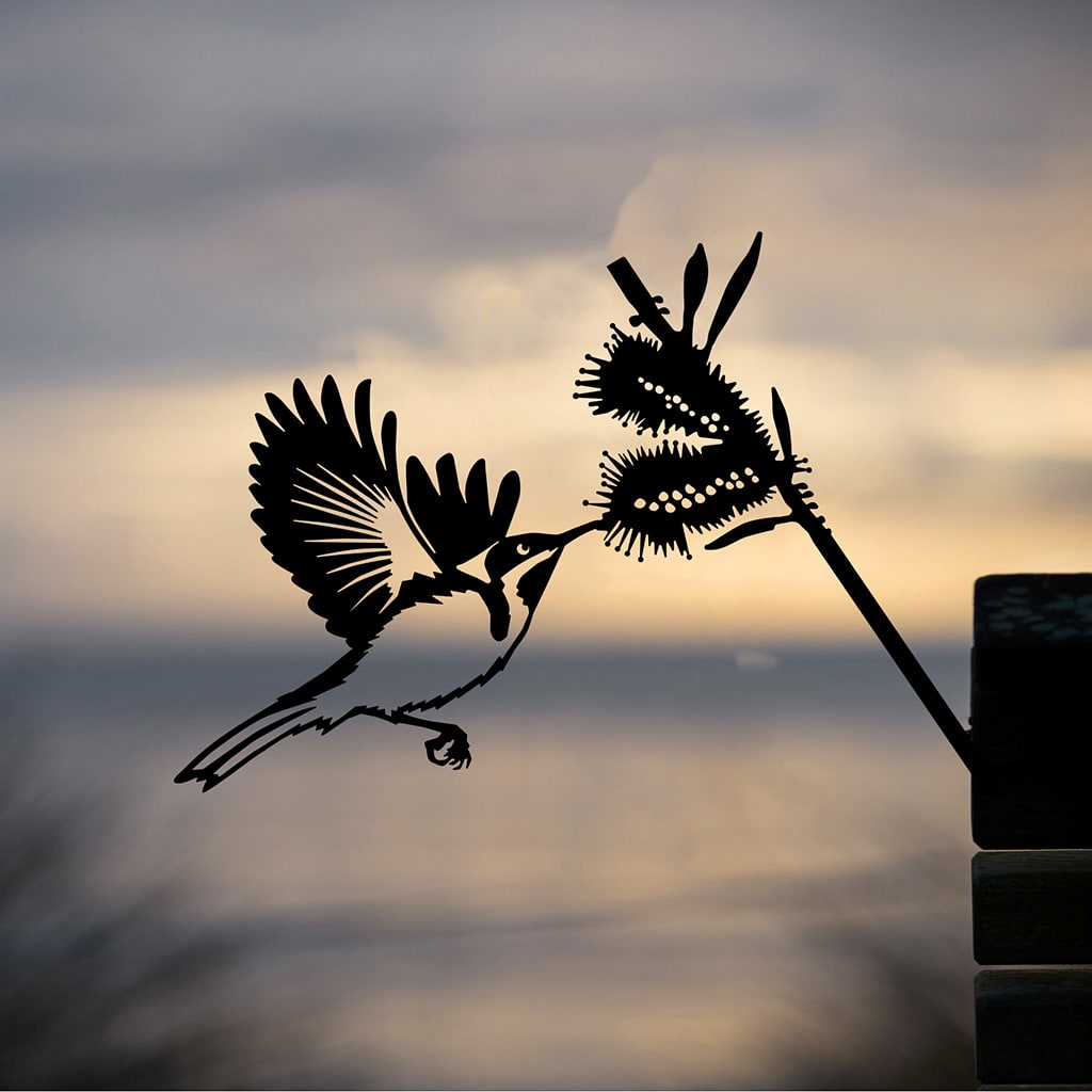 The Metalbird Australia Eastern Spinebill features a bird with open wings near a flower, creating striking metal yard art silhouetted against a soft, blurry sunset sky and ocean backdrop.