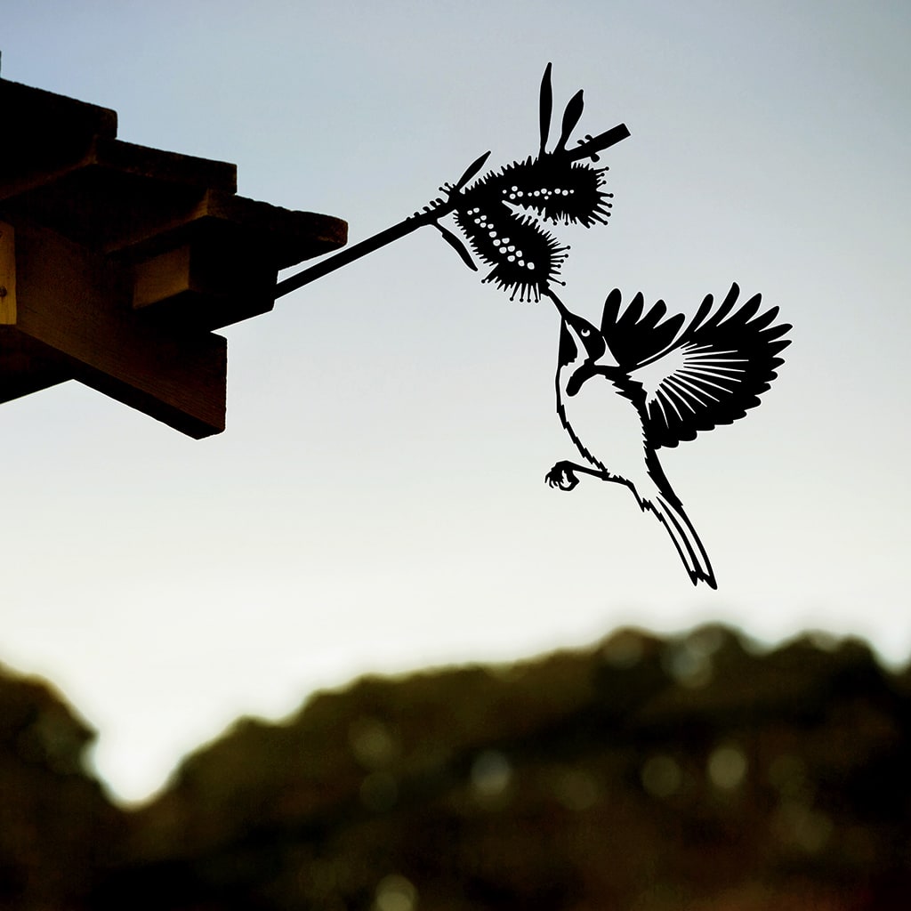 The Metalbird Australia "Eastern Spinebill" is a black metal silhouette of a bird feeding from a flower, mounted on the roof edge to create striking yard art against a blurred outdoor backdrop with cloudy sky and trees.