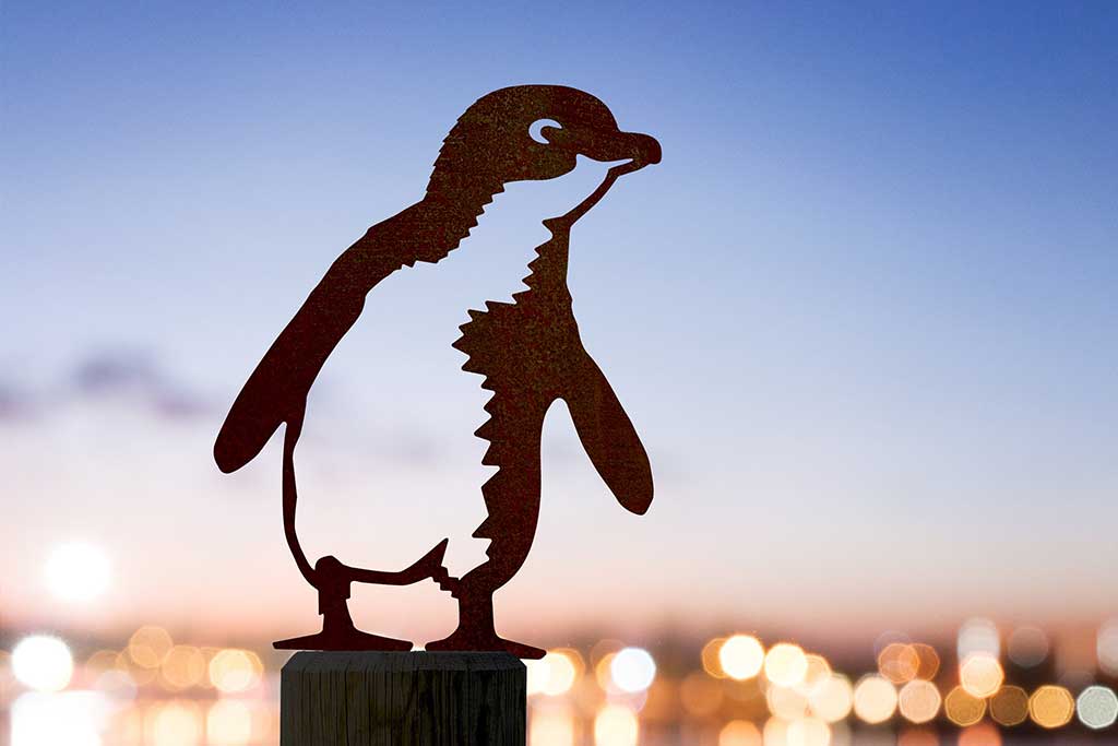 The Metalbird Australia Penguin is a premium metal silhouette on a post, creating unique yard art with city lights softly glowing in the dusk background.