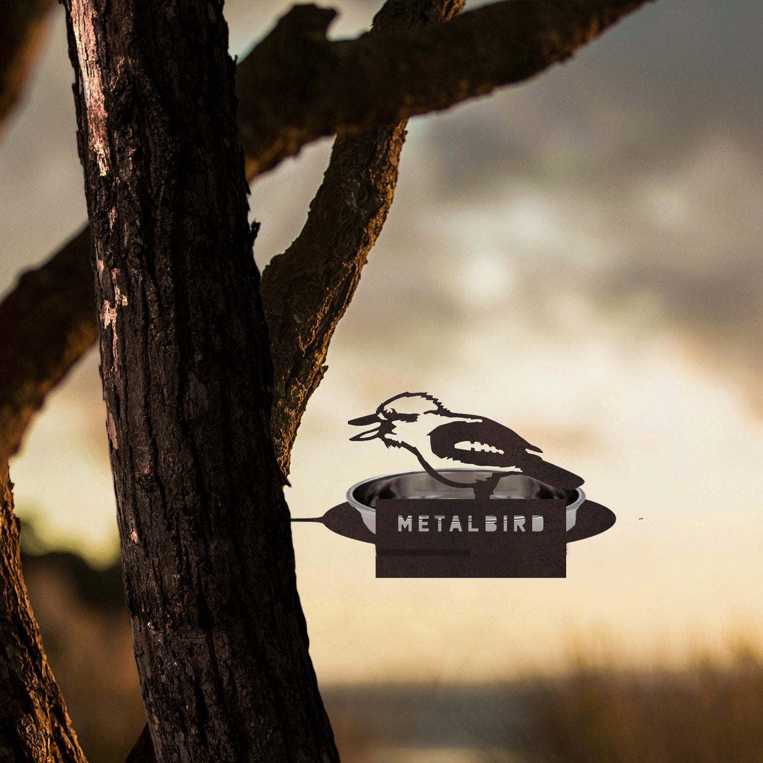 A premium Metalbird Australia Water Bowl Feeder, crafted from corten steel with "METALBIRD" cut into it, features a metal bird silhouette perched on a feeder attached to a tree trunk, set against a soft-focus outdoor sunset background.
