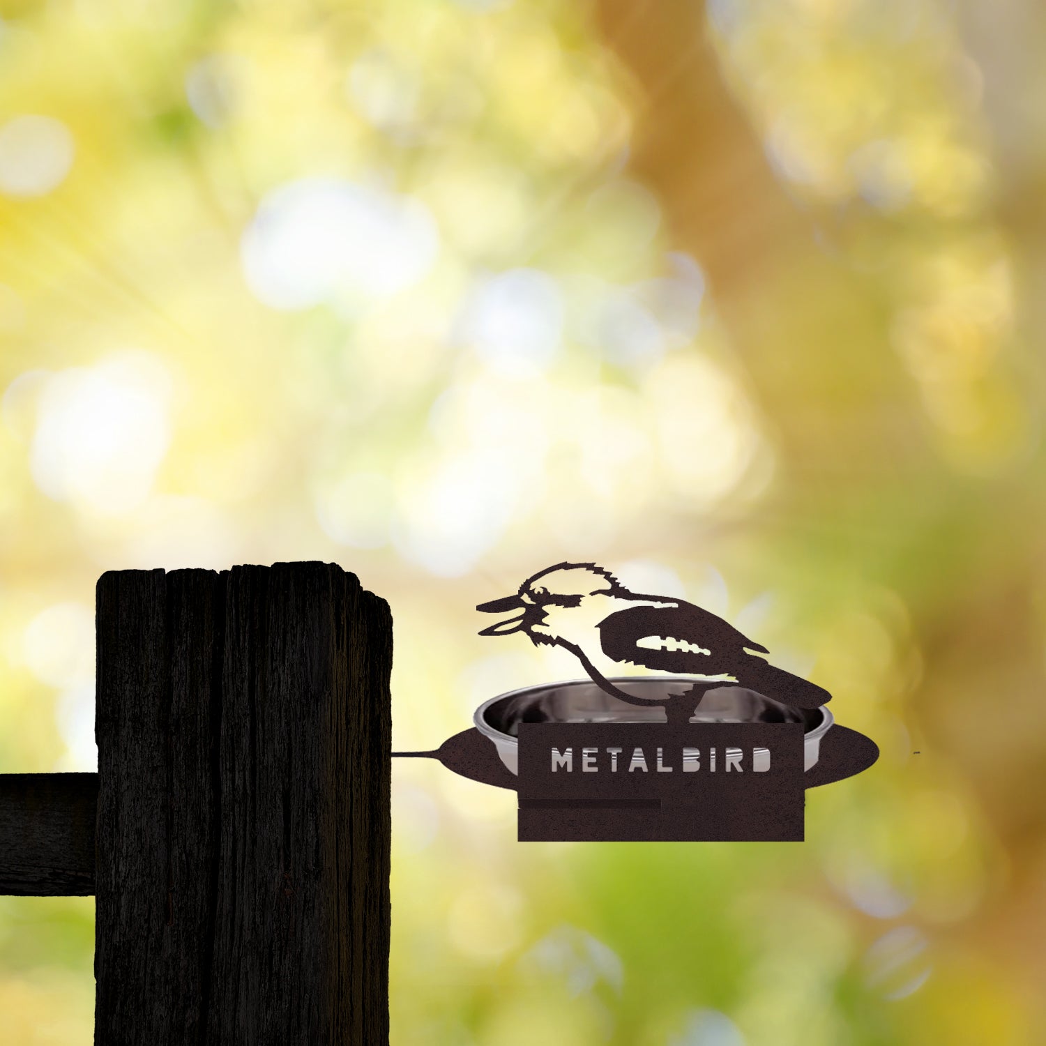 A premium Kookaburra silhouette from Metalbird Australia sits on a Water Bowl Feeder, mounted on a wooden post against a sunlit, tree-filled background. Crafted from durable corten steel for lasting beauty.