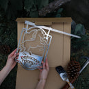 A person holds Metalbird Australia's THE PRANCING PONY™ metal sign—depicting a rearing horse and text—above an open box. Pinecones, tools, and a paintbrush lie nearby on grass, making it perfect as unique yard art or a thoughtful gift.