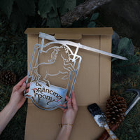 A person holds Metalbird Australia's THE PRANCING PONY™ metal sign—depicting a rearing horse and text—above an open box. Pinecones, tools, and a paintbrush lie nearby on grass, making it perfect as unique yard art or a thoughtful gift.