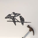 The Pair of Lorikeets by Metalbird Australia features a corten steel silhouette of two birds on a branch, mounted on a white wall with a hammer below—suggesting recent installation. A charming and thoughtful gift idea.