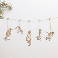 Five festive Winged Hangers from Metalbird Australia hang on a string with clothespins against a white background, a pine branch in the top left. Crafted from sustainably grown wood—perfect as wooden bird decorations.