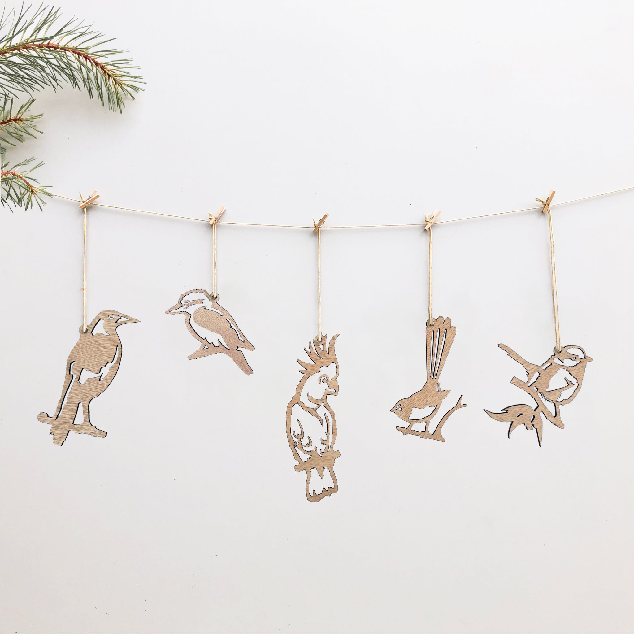 Five festive Winged Hangers from Metalbird Australia hang on a string with clothespins against a white background, a pine branch in the top left. Crafted from sustainably grown wood—perfect as wooden bird decorations.