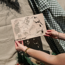 Hands hold a laser-cut metal sheet labeled Metalbird Australia, featuring five Winged Hangers festive decorations. A green checkered cloth and beige fabric background are visible.