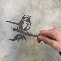 A hand holds the UPSELL Mini Blue Wren, a small corten steel bird silhouette perched on a branch, standing out against the gray concrete background.