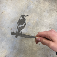 A hand holds the Mini Magpie by Metalbird Australia, a premium paper cutout of a bird on a branch against a plain, light background—perfect as a unique gift.