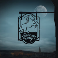 A THE PRANCING PONY™ metal silhouette sign by Metalbird Australia features a rearing horse, hanging against a twilight sky with a full moon and distant village in the background.