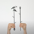 Two hands hold up Metalbird Australia's Kookaburra Plant Stake—a black corten steel piece featuring a bird on a leafy branch—and a simple branch yard art stake, both displayed against a plain white background.