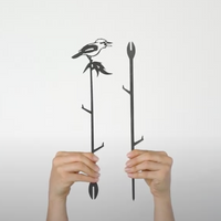 Two hands hold up Metalbird Australia's Kookaburra Plant Stake—a black corten steel piece featuring a bird on a leafy branch—and a simple branch yard art stake, both displayed against a plain white background.