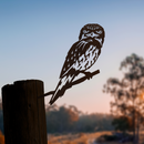 A corten steel silhouette of the Tawny Frogmouth by Metalbird Australia perches on a wooden post, framed by blurred trees and a gentle blue dusk sky in the background.