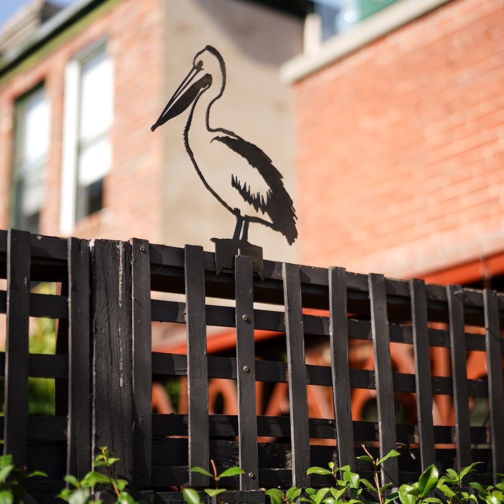 The Metalbird Australia Pelican is a premium corten steel silhouette, shown perched on a wooden lattice fence with a brick building and lush green foliage in the background.