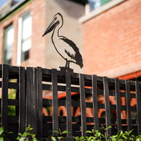 The Metalbird Australia Pelican is a premium corten steel silhouette, shown perched on a wooden lattice fence with a brick building and lush green foliage in the background.